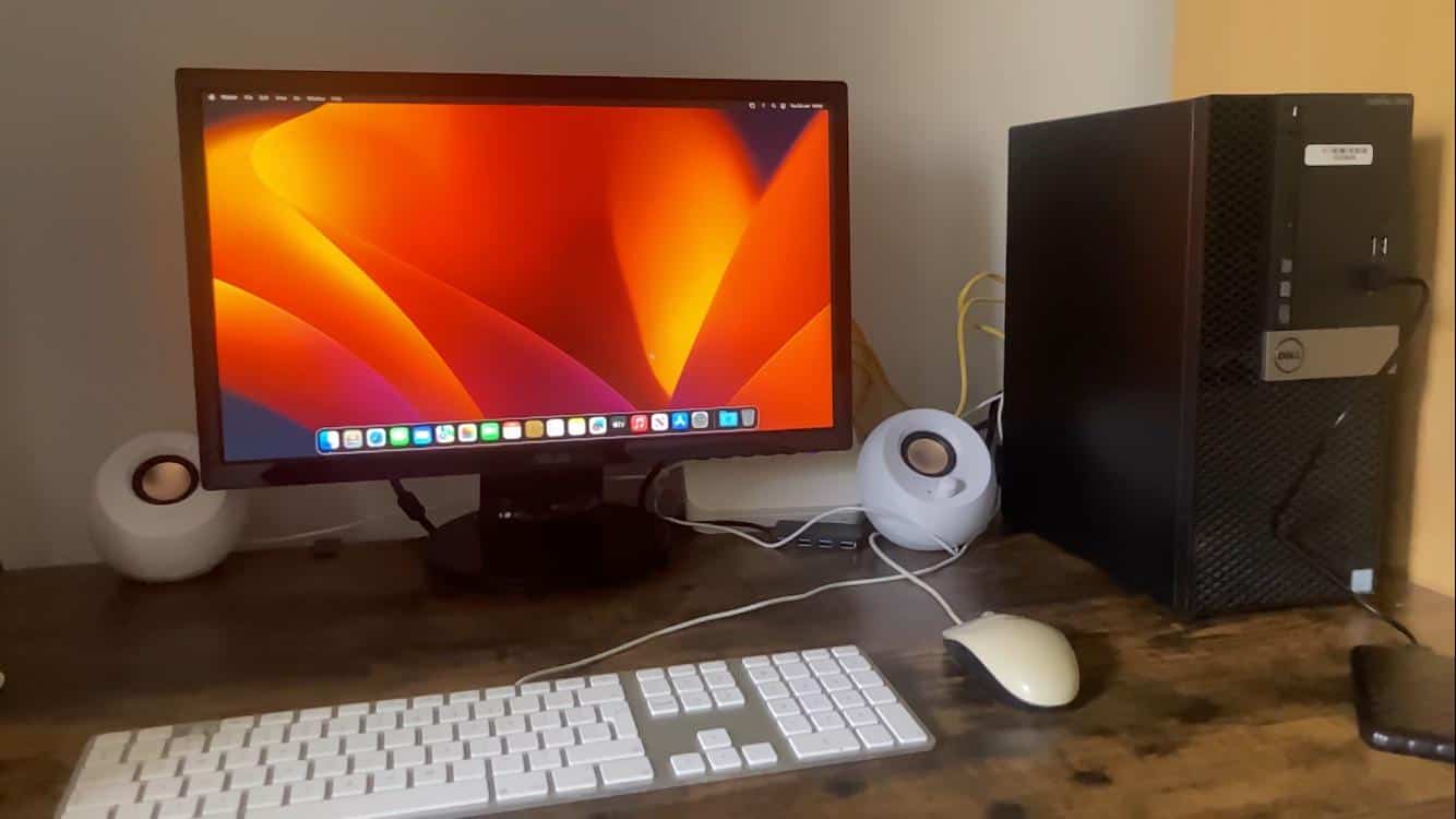 Monitor showing a macOS start screen with a Dell desktop computer to its right, along with mouse, keyboard and other peripherals over a table